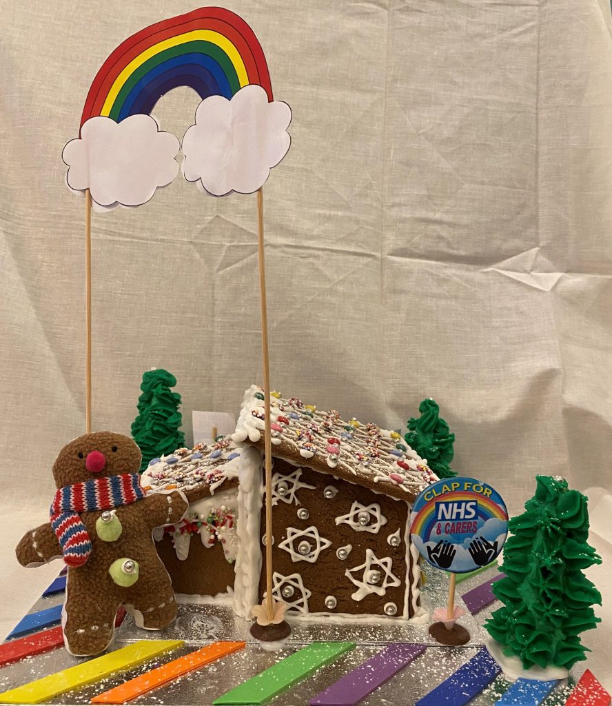 Gingerbread house surrounded with rainbow icing strips. Fir trees made from green icing stand in 3 of the corners. In the third corner stands a gingerbread person soft toy. The house is overlooked by a paper rainbow held up with thin wooden sticks.